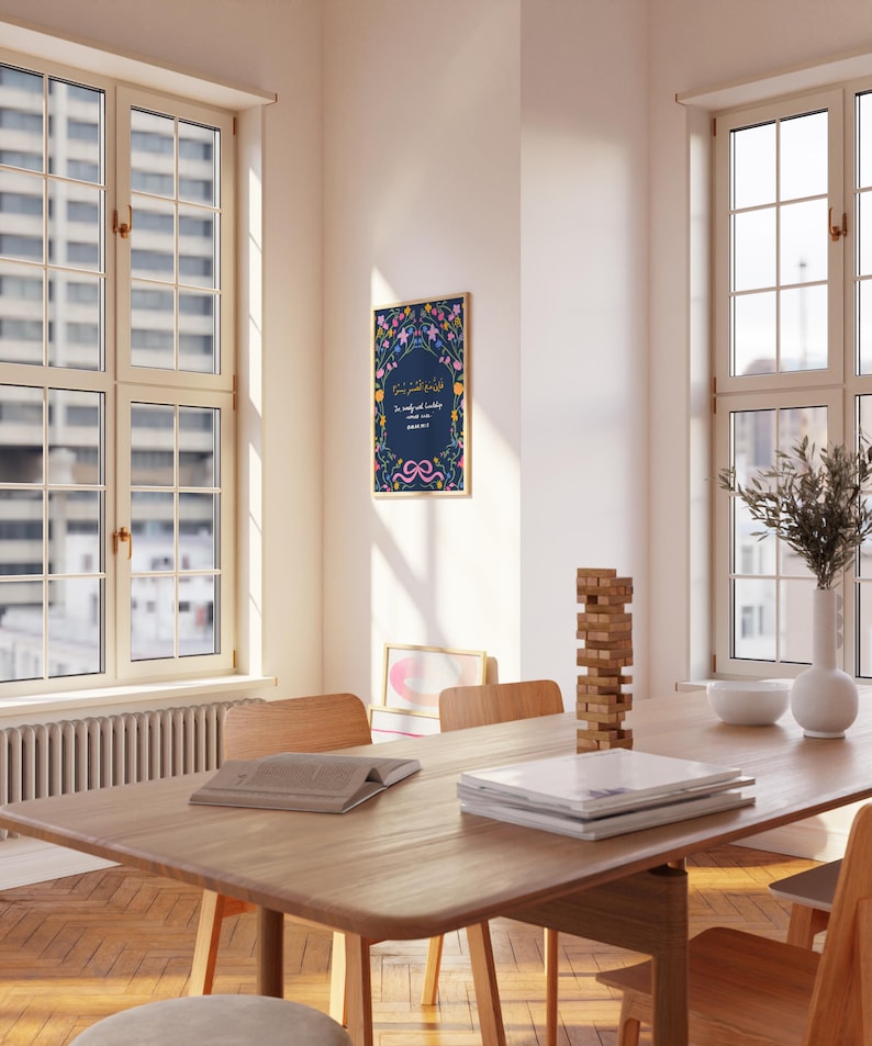 May include: A framed art print with Arabic calligraphy and floral accents hangs on a white wall. The print features a dark blue background with colorful flowers and text. The room has a wooden table, chairs, and large windows.