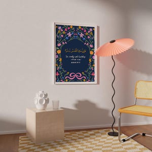 May include: A framed art print with a navy blue background and floral border. The print features Arabic script and English text, with a pink ribbon design at the bottom. The frame is white, and the print is hanging on a white wall.