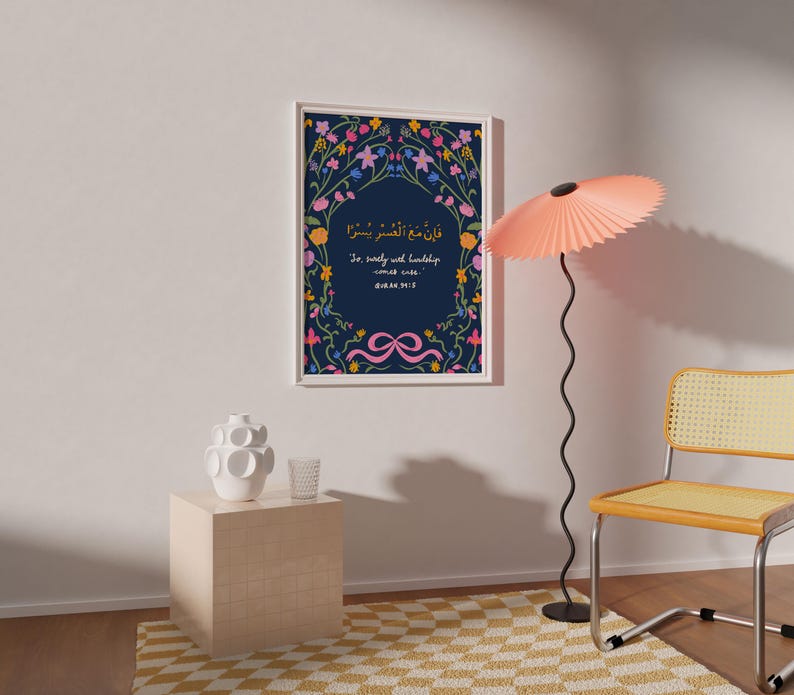 May include: A framed art print with a navy blue background and floral border. The print features Arabic script and English text, with a pink ribbon design at the bottom. The frame is white, and the print is hanging on a white wall.