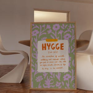 May include: A framed print with the word "HYGGE" in orange, with a definition below. The background is a green floral pattern with purple flowers. The frame is light brown.