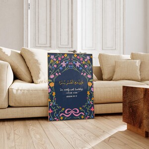 May include: Framed art print featuring a navy blue background with floral and botanical illustrations in pink, yellow, and blue. The print includes Arabic script and English text: "So, surely with hardship comes ease." The artwork is displayed on a wooden floor.