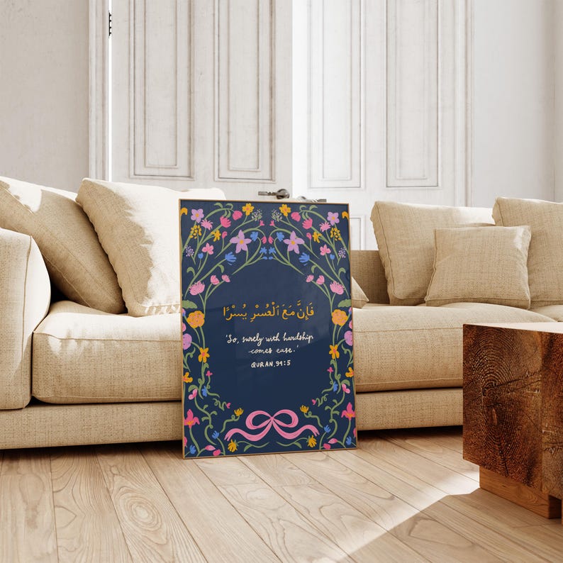 May include: Framed art print featuring a navy blue background with floral and botanical illustrations in pink, yellow, and blue. The print includes Arabic script and English text: "So, surely with hardship comes ease." The artwork is displayed on a wooden floor.