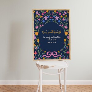 May include: A framed art print featuring a navy blue background with floral accents in pink, yellow, and blue. The print includes Arabic script and English text: "So, surely with hardship comes ease." A pink ribbon design is at the bottom.