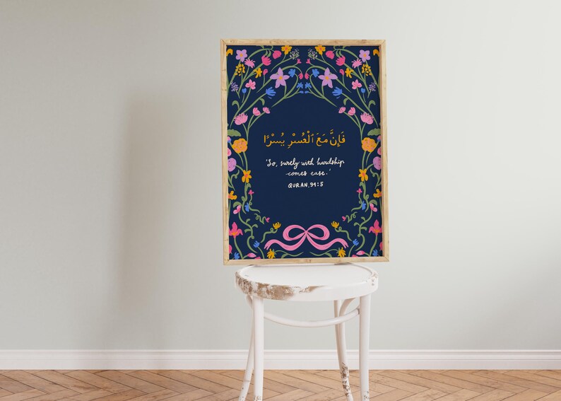 May include: A framed art print featuring a navy blue background with floral accents in pink, yellow, and blue. The print includes Arabic script and English text: "So, surely with hardship comes ease." A pink ribbon design is at the bottom.