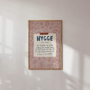 Op de afbeelding: Een ingelijste print met het woord "HYGGE" in marineblauw op crèmekleurig papier, geplakt op een roze bloemenachtergrond. De definitie van "hygge" staat eronder. De lijst is lichtbruin.