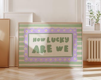 May include: A framed art print with the text "How Lucky Are We" in a playful font. The artwork features a light green and lavender color scheme with a floral border and striped background. The print is displayed in a room with natural light.