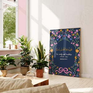 May include: A framed art print featuring a navy blue background with floral accents in pink, yellow, and blue. The print includes Arabic script and the English phrase "So, surely with hardship comes ease." The artwork is displayed in a well-lit room with potted plants.