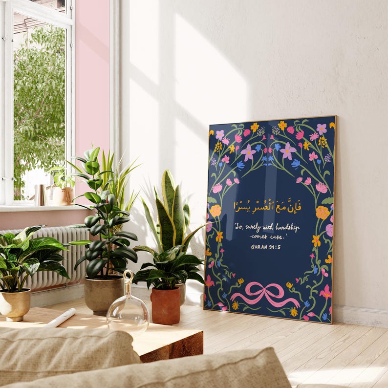 May include: A framed art print featuring a navy blue background with floral accents in pink, yellow, and blue. The print includes Arabic script and the English phrase "So, surely with hardship comes ease." The artwork is displayed in a well-lit room with potted plants.