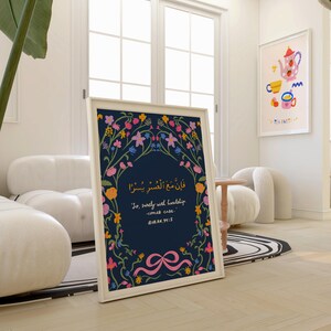 May include: A framed art print with a navy blue background and floral border. The print features Arabic script and English text: "So, surely with hardship comes ease." A pink ribbon design is at the bottom. A second framed print with a tea party theme is visible.