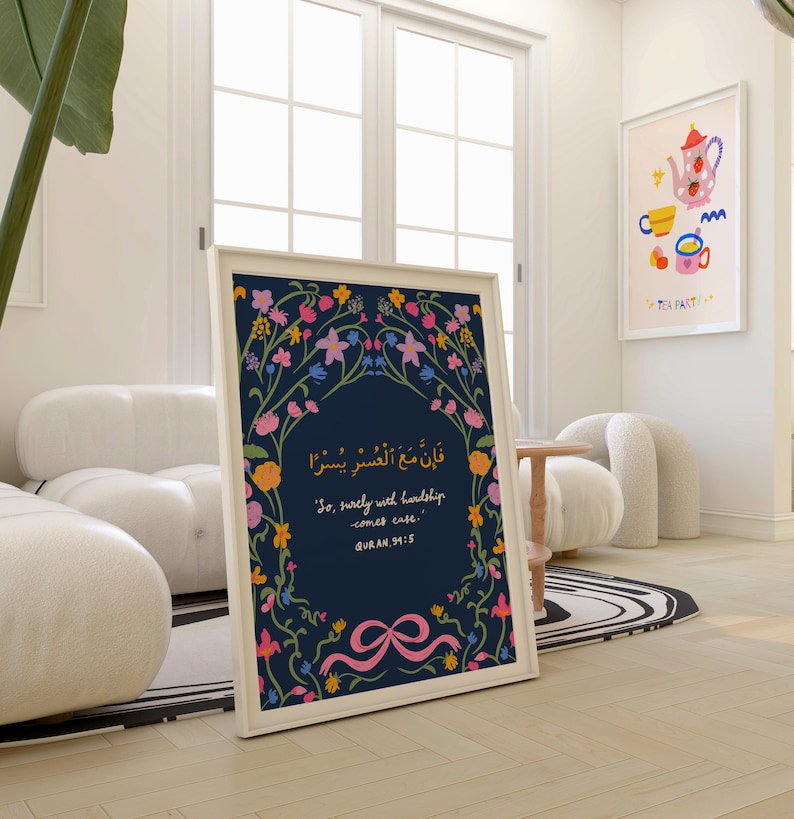May include: A framed art print with a navy blue background and floral border. The print features Arabic script and English text: "So, surely with hardship comes ease." A pink ribbon design is at the bottom. A second framed print with a tea party theme is visible.