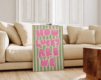 May include: A framed art print with the text "HOW LUCKY ARE WE" in pink bubble letters on a green and white striped background. The artwork is displayed on a wooden floor, leaning against a beige sofa.