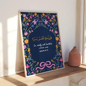 May include: A framed art print featuring a navy blue background with floral and vine accents in pink, yellow, and blue. The print includes Arabic script and the English phrase "So, surely with hardship comes ease." A pink ribbon is at the bottom.