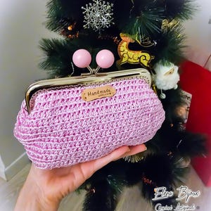 Può includere: Un porta monete rosa lavorato a uncinetto con una cornice in metallo dorato e due bottoni in legno rosa. Il porta monete ha un'etichetta in pelle marrone che dice "Hand made with love".