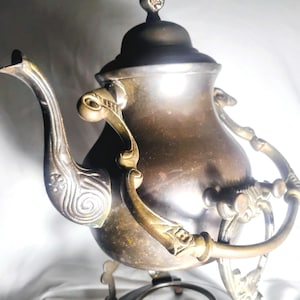 May include: Antique silver-toned tea kettle with ornate detailing. The kettle features a curved spout, decorative handles, and a domed lid with a finial. It is supported by a decorative stand with a circular base, showcasing a vintage aesthetic.