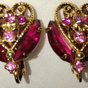 May include: A pair of vintage clip-on earrings. Each earring features a heart-shaped design with a gold-tone filigree frame. The earrings are adorned with red and pink gemstones, creating a glamorous and eye-catching accessory.