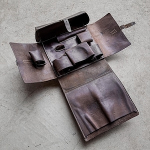 May include: A brown leather tool roll, open to display its interior. It features various pockets and compartments for tools. The leather has a distressed look, with visible stitching and a buckle fastening. The tool roll is laid flat.