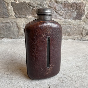 May include: A vintage brown leather flask with a metal cap. The flask has a rectangular shape with rounded edges and a vertical indentation. The leather shows signs of wear, giving it a rustic appearance. The cap is made of a silver-colored metal.