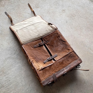 Puede incluir: Un bolso de cuero marrón vintage con una textura similar a la piel en la parte delantera y dos bolsillos delanteros. El bolso tiene correas y hebillas de cuero, y un interior de tela de color claro. El bolso está abierto, revelando el interior.