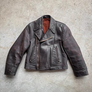May include: A brown leather jacket with an asymmetrical zip. It has a notched collar, two zipped pockets, and a reddish-brown lining. The leather has a distressed look, giving it a vintage feel. The jacket's design is classic and timeless.