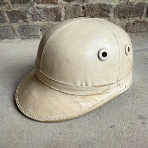 May include: A white leather helmet with a wide brim and two small holes on the top. The helmet has a worn, vintage look.