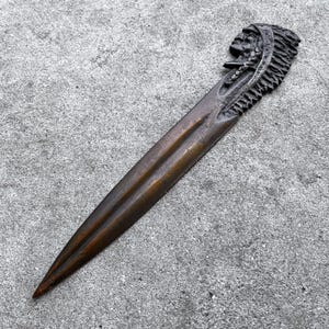 May include: A bronze letter opener with a Native American chief's head on the handle. The opener has a long, pointed blade and is made of a dark, metallic material. The handle is intricately detailed.