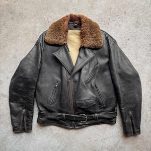 May include: A black leather jacket with a brown shearling collar. The jacket has a belted waist, multiple zipped pockets, and a front zip closure. The jacket has a vintage, worn appearance.