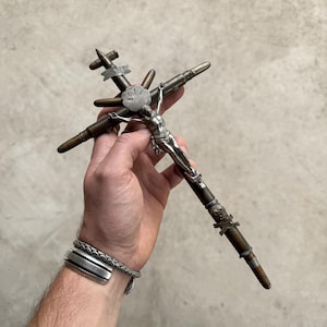 May include: A crucifix constructed from metallic bullet casings and components, featuring a silver-toned figure of Jesus at the center. The cross includes a small metal airplane silhouette at the top, a flower-shaped emblem with the text 1914, and a skull and crossbones detail on the lower vertical shaft.