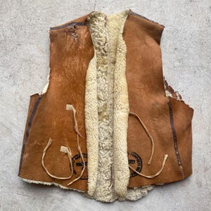 Puede incluir: Un chaleco de cuero marrón vintage con forro de borrego color crema. El chaleco presenta signos de desgaste, con agujeros y bordes deshilachados. Tiene cierre frontal y detalles de lazos. El diseño sugiere un estilo rústico.