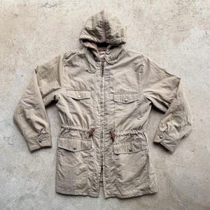 Könnte beinhalten: Eine beige Kapuzenjacke mit durchgehendem Frontreißverschluss, zwei Brusttaschen mit Druckknopfverschluss und zwei unteren Taschen mit Druckknopfverschluss. Die Taille verfügt über einen Kordelzug mit braunen Knebelknöpfen an den Enden.