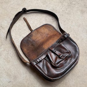 Könnte beinhalten: Eine dunkelbraune Umhängetasche aus Leder mit einer großen Umschlagklappe, einem verstellbaren Schulterriemen und einer Vordertasche, die mit einer Schnalle gesichert ist. Die Innenseite der Klappe zeigt eine abgenutzte, hellbraune Textur.