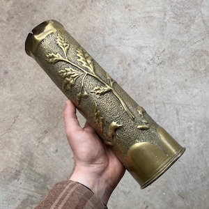 Puede incluir: Objeto cilíndrico de latón con diseño en relieve de hojas de roble y bellotas. El objeto tiene una superficie texturizada y una abertura ligeramente ensanchada. El color general es un dorado antiguo cálido.