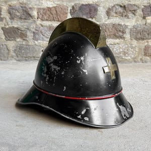 Pode incluir: Um capacete de bombeiro de metal preto com uma crista de latão no topo, um emblema de cruz de latão na frente e uma fina faixa horizontal vermelha ao redor da base. O capacete apresenta sinais de desgaste com lascas de tinta visíveis.