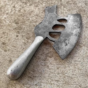 May include: A vintage metal axe with a silver handle and a weathered, gray blade. The axe head features three circular cutouts. The tool is designed for chopping or splitting wood and is set against a textured, concrete surface.