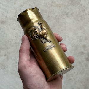 May include: A cylindrical brass artillery shell casing featuring a raised relief of a rooster and a small banner with the text Verdun. The metal surface has a polished, weathered patina with visible metallic textures and a flared top rim.