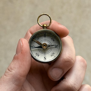 Könnte beinhalten: Ein kleiner, runder Kompass aus Metall mit einem messingfarbenen Gehäuse und einem oberen Aufhängering. Das weiße Zifferblatt zeigt die Himmelsrichtungen N, S, W und SW, wobei die zentrale Nadel einen sichtbaren braunen Oxidationsfleck aufweist.