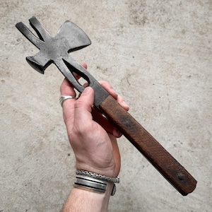 Puede incluir: Una herramienta multiusos vintage con martillo, hacha y otras funciones. La cabeza de metal tiene un aspecto oscuro y envejecido, y el mango es de madera teñida. La herramienta se sostiene en una mano sobre un fondo neutro.