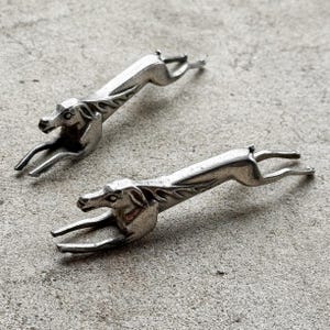 Puede incluir: Dos figuras de perro de metal plateado en pose de carrera. Los perros tienen detalles precisos, incluyendo orejas puntiagudas y patas delgadas. Las figuras están colocadas sobre una superficie gris texturizada.