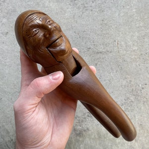 May include: A wooden nutcracker carved in the shape of a person's head with a wide smile. The nutcracker is brown and has a hinged jaw that opens and closes to crack nuts.