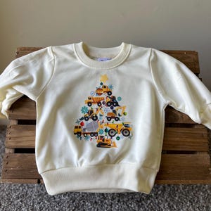 May include: A cream-coloured sweatshirt with a Christmas tree design featuring construction vehicles. The tree is decorated with yellow lights, red baubles, and snowflakes.