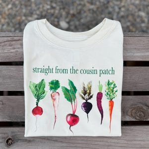 May include: Cream-colored t-shirt featuring a watercolor illustration of various root vegetables, including radishes, carrots, and beets. The text "straight from the cousin patch" is printed in green above the vegetables.