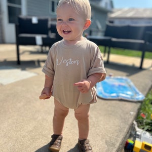 Puede incluir: Un body de bebé color beige con el nombre "Veston" en escritura blanca. El body tiene mangas cortas y cuello redondo. El niño lleva zapatos de cuero marrón. El fondo incluye una casa, un patio y un cielo soleado.