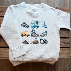 May include: A light grey sweatshirt featuring a graphic print of various construction vehicles, including a crane, bulldozer, dumper truck, and excavator. The vehicles are yellow and orange with blue accents.