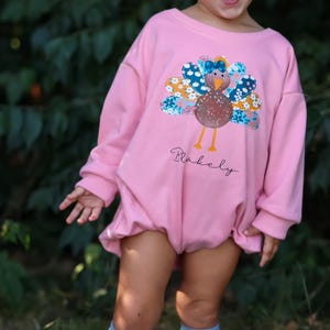 May include: A pink long-sleeved onesie with a colorful turkey applique and the name "Blakeely" written in cursive. The onesie is worn by a young child with blue socks and brown shoes.