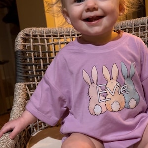 May include: A young child wearing a lavender t-shirt with a graphic of three pastel bunnies and the name "Evie" printed on the front. The child is sitting on a wicker chair.