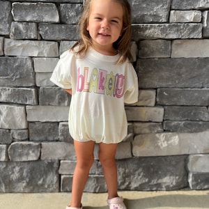 May include: A young child wearing a white t-shirt with the name "BLAKELEY" written in colorful letters. The child is wearing a purple bow in their hair and pink and purple shoes.