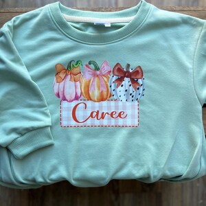 May include: A light green sweatshirt featuring a pumpkin design with bows and the name "Caree" on a gingham background. The pumpkins are in shades of pink, orange, and white with decorative bows. The sweatshirt has a crew neck and long sleeves.