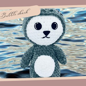 May include: A crocheted plush toy shaped like a shark with a white belly and large black eyes. The toy is blue-grey and is photographed against a blue and white water background. The text "Bubble shark" is written in cursive at the top of the image.