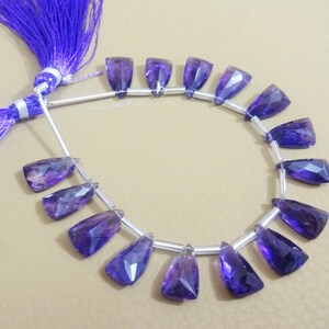 May include: A strand of purple amethyst gemstone beads, each bead is faceted and shaped like a teardrop. The beads are strung on a silver wire and have a purple tassel at each end.