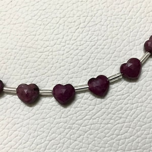 Pode incluir: Um fio de contas de pedras preciosas em forma de coração. As contas são de cor roxa escura e são enfiadas em um fio de prata.