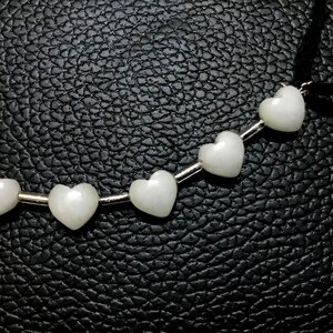 May include: A white heart-shaped bead necklace with a black cord. The necklace is made of five heart-shaped beads connected by silver metal.